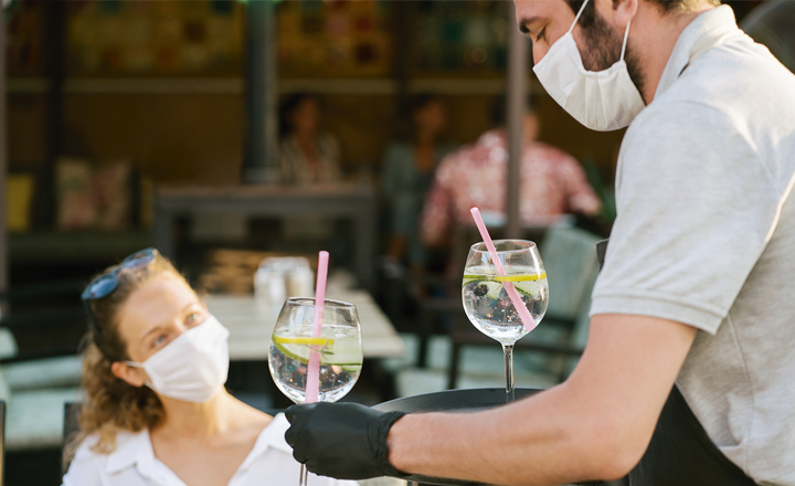 Waiter serving woman a drink outside, both in masks